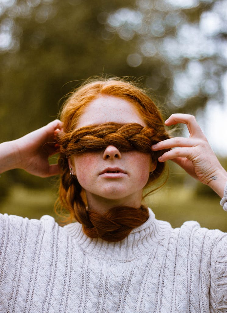 A young woman with red braided hair covers her eyes in an outdoor setting, exuding a mysterious vibe.