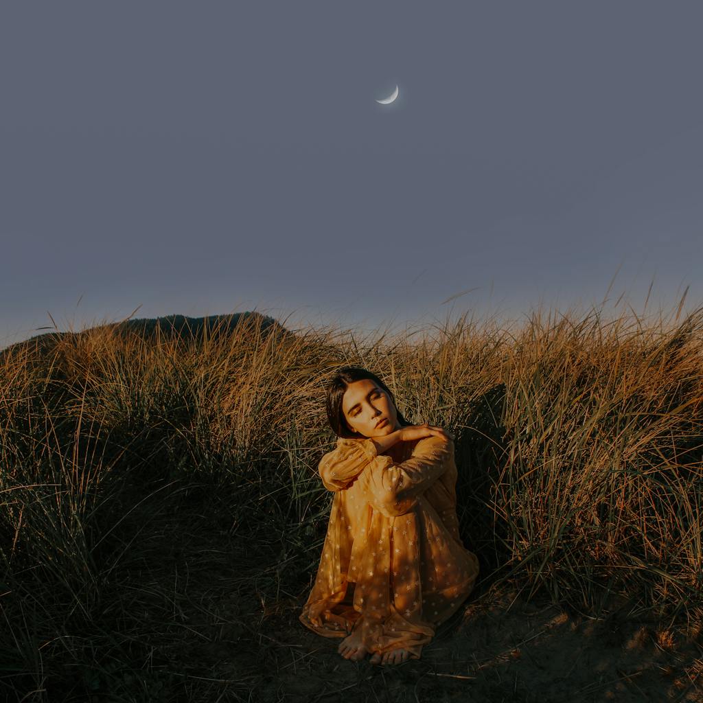 Woman in an orange dress sitting in grass, dreaming under a crescent moon at Cannon Beach.